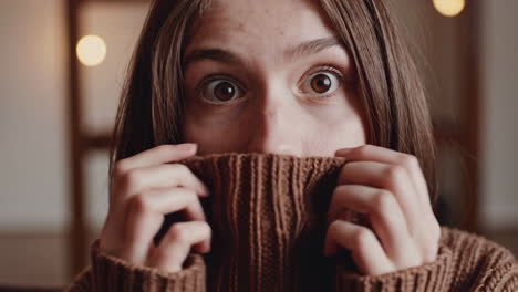young woman with wide eyes covering her face with a brown sweater