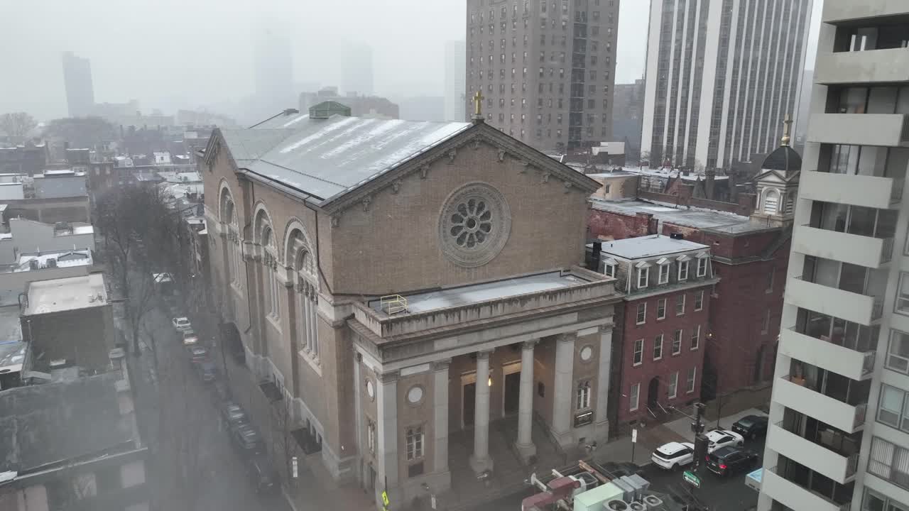 Chiesa cattolica di San Patrizio a Filadelfia durante una fitta nevicata. La storica facciata in pietra e il rosone spiccano tra gli edifici a più piani circostanti e i tetti coperti di neve. Vista aerea.