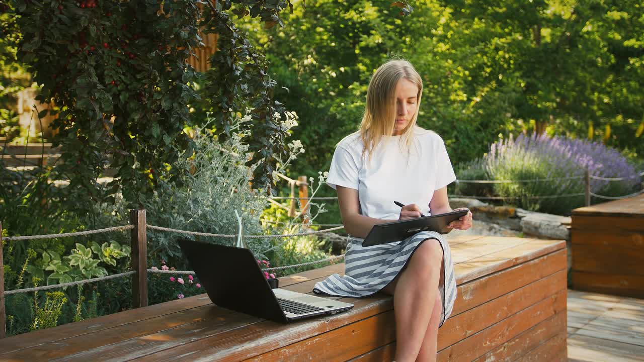 Young woman is sitting on a bench of city park and drawing on a digital tablet, using laptop. Summer sunny day. Concept of freelance