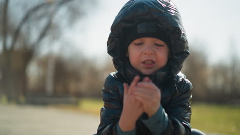a young boy wearing a shiny black jacket and head warmer, with his mouth slightly open as he rubs his hands together, with a blur background of trees and a house