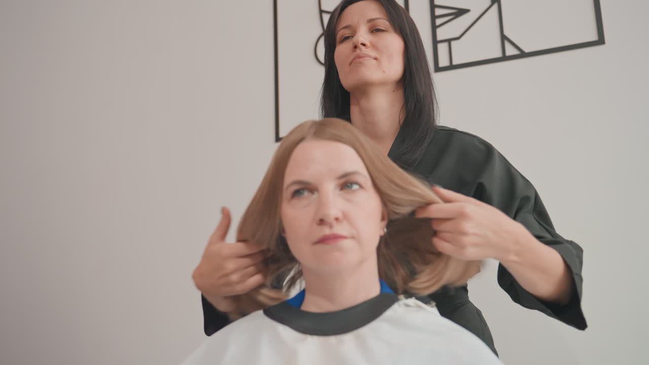 Una clienta en la silla del salón recibe un corte de pelo y un peinado de un estilista atento; capa sobre los hombros, peinando y alisando mechones para crear un acabado liso y elegante; interior moderno y minimalista.