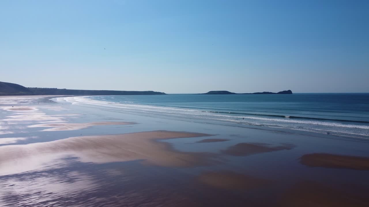 Establishing Aerial View of Rhossili Bay with Worm's Head Causeway Landmark Headland with Sun Reflection and Clear Blue Sky with Golden Sands 4K