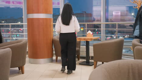 confident woman walks toward restaurant table, places dinner down, then relaxes into cushioned chair with legs crossed while calmly admiring illuminated cityscape through wide glass window at night
