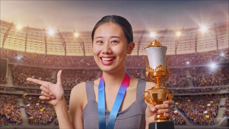close up of asian woman with a gold medal holding a gold trophy in her hands, smiling, and pointing to side at the stadium