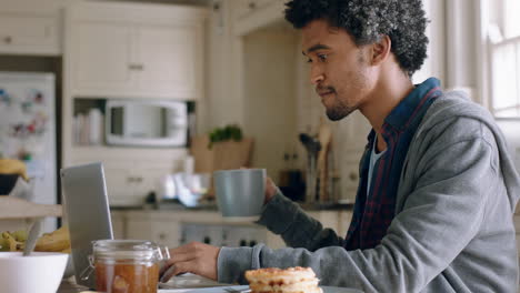 happy mixed race man using laptop computer working at home in kitchen typing email drinking coffee enjoying modern wireless communication technology