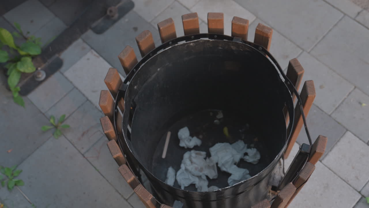 Person Drops Tissue Into Bin, Overhead Shot Of Hand Releasing Crumpled Tissues Into Wooden Waste Container On Tiled Pavement, Nearby Plant And Green Edging Visible, Deliberate Ritualized Motion