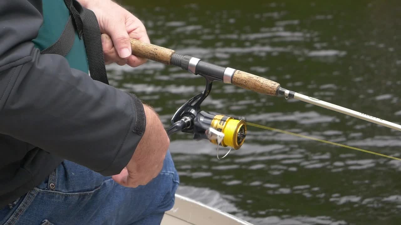 Slow-motion close-up of fisherman’s hands reeling fishing line with water and jeans visible in background.