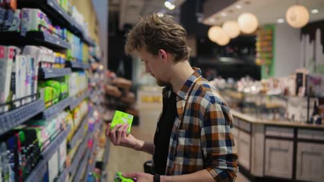 Caucasian-young-man-holding-feminine-pads-in-the-mall-and-reading-the-label,-grimacing.-Concept-of-shopping-and-choices.-Side-view.-Slow-motion