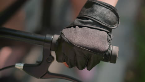 close-up of individual gripping bike handlebar with gloved hand, holding brake lever, sunlight reflects subtly off the leather glove, background shows a soft blur of greenery