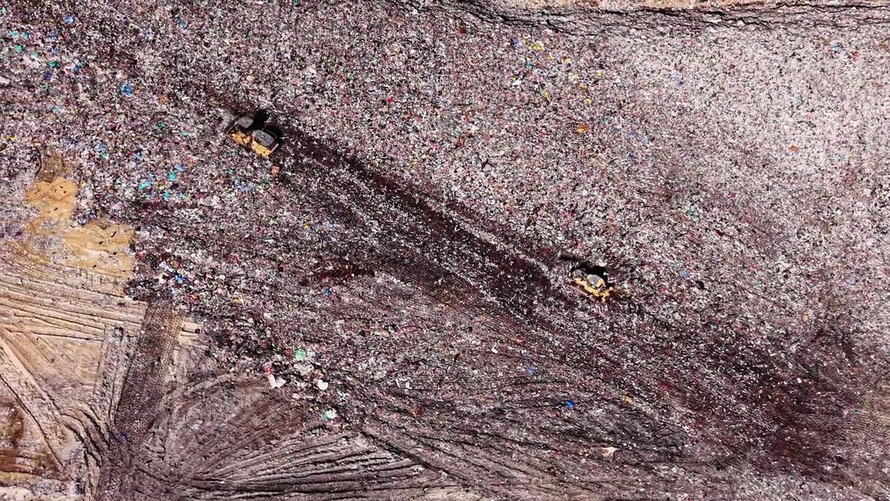 Drohnenaufnahme aus großer Höhe, die detailliert zeigt, wie schwere Fahrzeuge Spuren durch Tausende von Plastikverpackungen ziehen. Draufsicht auf Bulldozer, die Müll auf einer Deponie verschieben.
