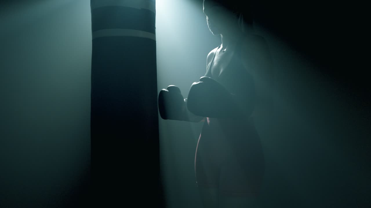 Donna bionda in piedi di profilo vicino a un sacco da boxe. Inquadrata dalle ginocchia in su, batte i guantoni da boxe tra loro, concentrata prima dell'allenamento. La telecamera si avvicina lentamente. Luce cinematografica in una palestra fumosa