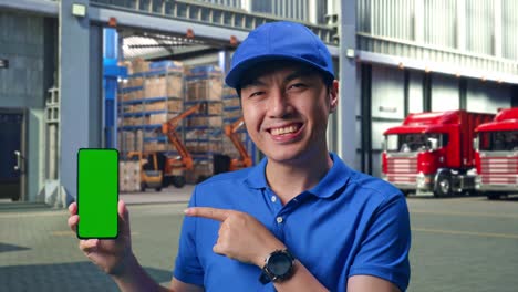 close up of asian male courier in blue uniform smiling and pointing to green screen smartphone while standing, outside of logistics distributions warehouse
