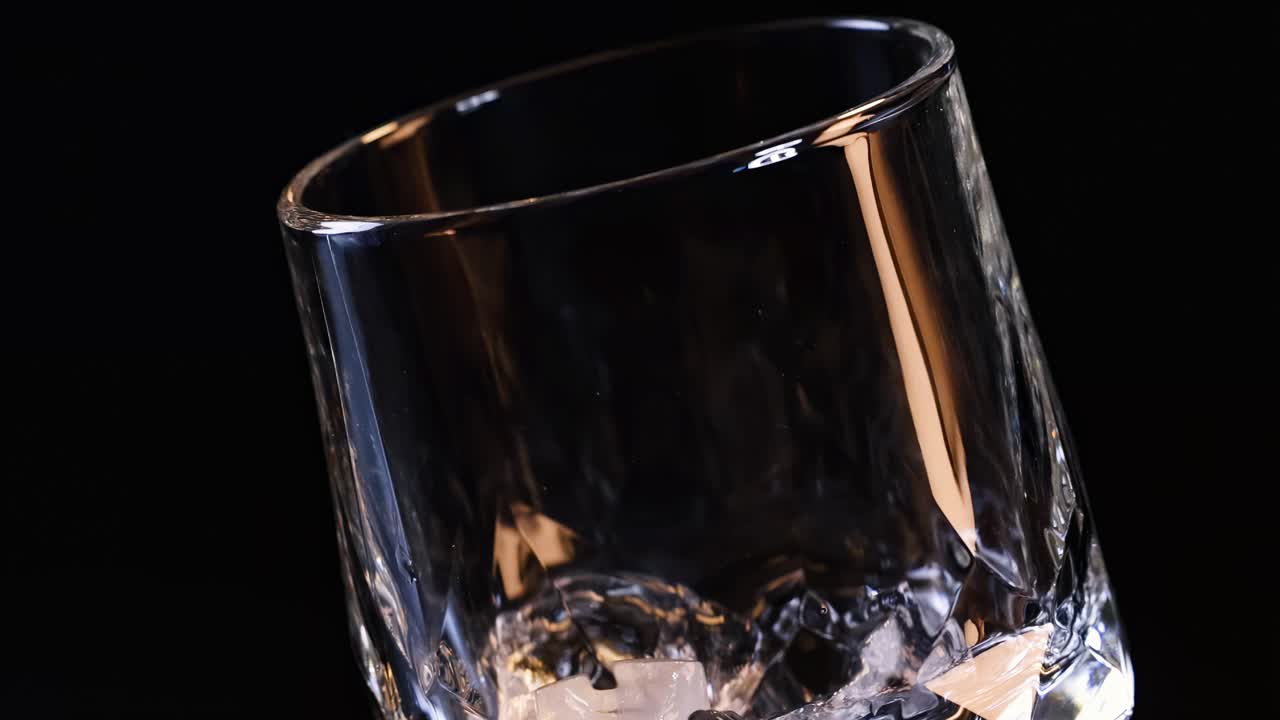 A hand places ice cubes into a tilted glass, set against a dark backdrop.