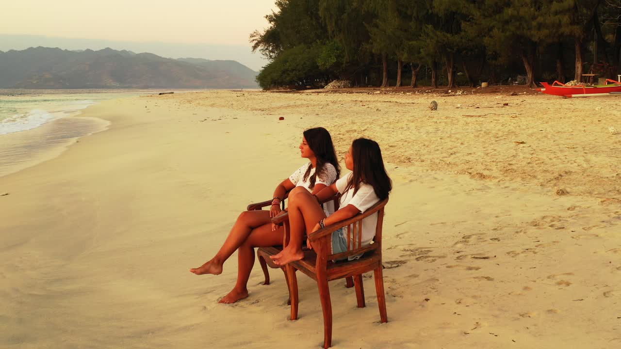 Two girls facing sea, sitting on chairs over white sand of exotic beach on a quiet sunset scenery of tropical island in Indonesia