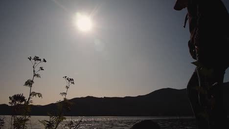 Silhouette-of-man-looking-at-view-with-sun-on-shimmering-lake-with-mountains-in-background