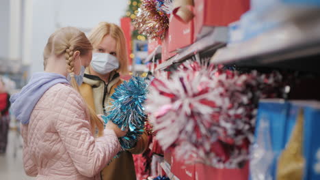 mom and daughter choose decorations for the christmas tree in the store. wear protective masks