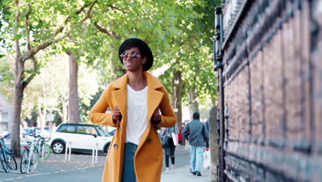 millennial black woman wearing an unbuttoned yellow coat and a hat walking down a city street past railings, selective focus