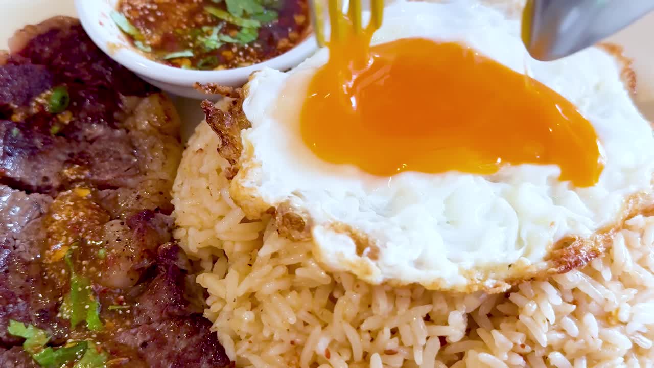 A close-up of a fried egg yolk drizzling over rice, accompanied by steak and sauce.