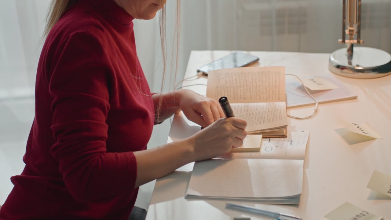 Lady in red turtleneck writes on sticky notes. Woman uses pens moves papers including labeled post it notes. Desk lamp white curtains window visible in background