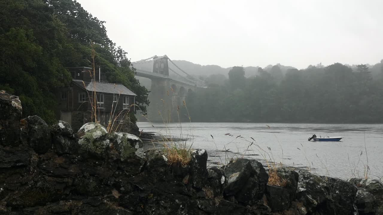 lluvia brumosa a través de la pintoresca casa de vacaciones en la orilla del río bajo el puente del estrecho de menai, anglesey, norte de gales