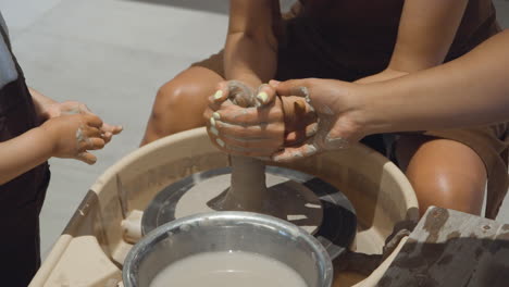 mother with daughter shaping mound of clay on potter wheel during pottery workshop with teacher's help - detailed close-up slow motion