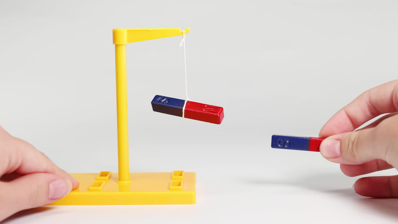 Hands manipulate bar magnets near a suspended magnet, demonstrating attraction and repulsion in a controlled environment