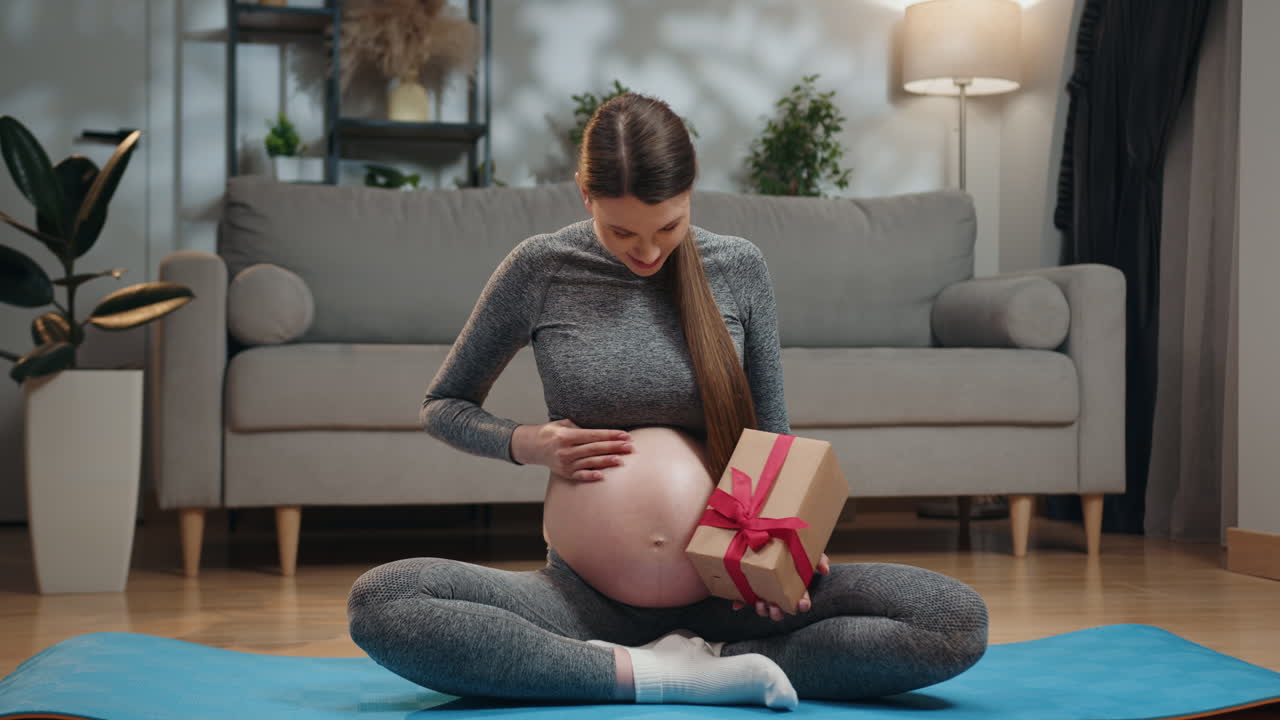 Pregnant woman in yoga attire sits crosslegged on mat holding gift box near belly indoors with sofa