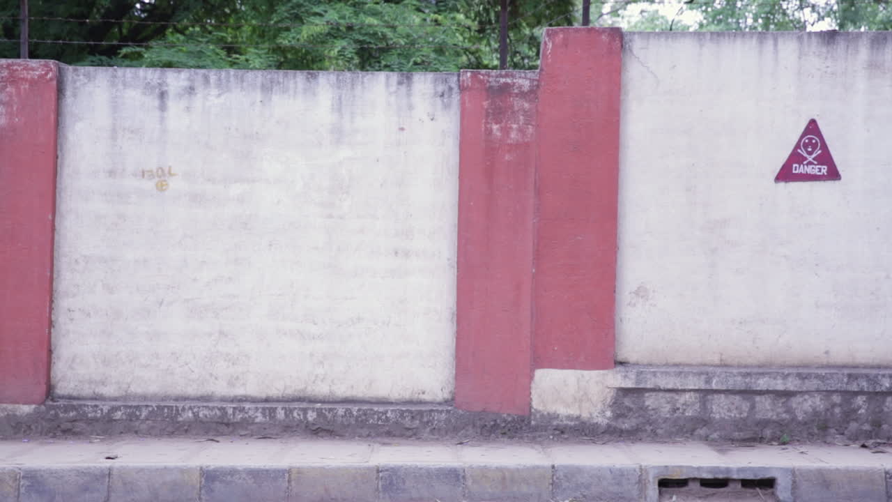 A compound wall in Bangalore marked with a danger sign and street traffic against a backdrop of green trees