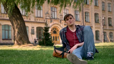 young handsome man is sitting on grass in park, listening music with earphones, relax concept, building on background