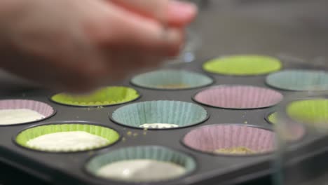 a woman scooping cheesecake cream filling with a spoon to fill mini muffin holders, which sit in a muffin pan, to make cheesecake cups