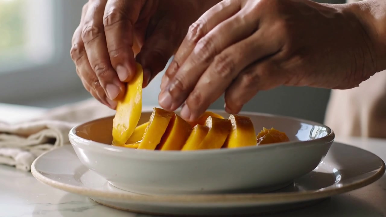 Mani che sistemano, disponendo a ventaglio fette di mango in cucina vicino alla finestra, con ciotola e polsino color cuoio, per servire