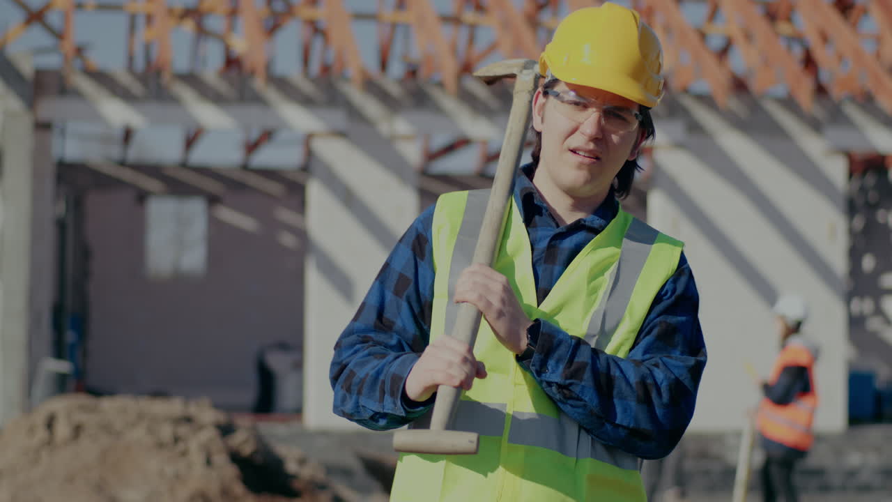 Portrait of confident young male worker wearing hardhat and reflective clothing carrying shovel on shoulder while standing at construction site