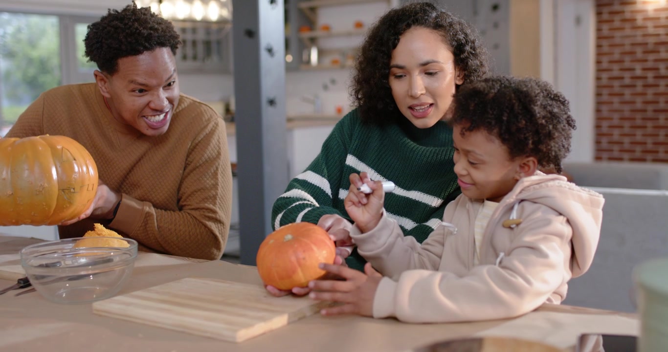 Famiglia afroamericana che decora zucche sull'isola della cucina, la madre usa un pennarello bianco e guida il bambino.