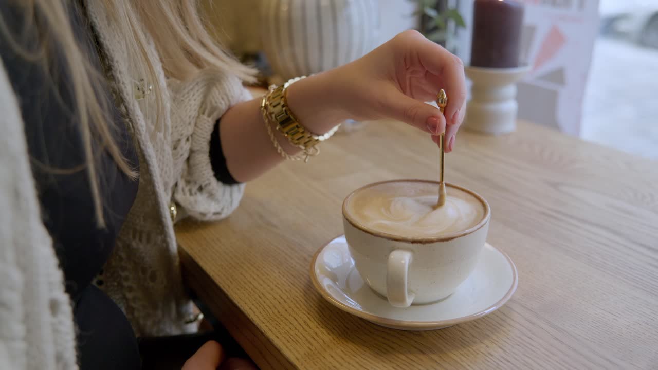 Female slowly stirring cup of coffee with golden spoon in cafe