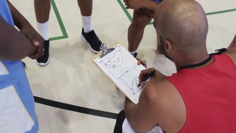 diverse male basketball team and coach with clipboard in discussion at indoor court, in slow motion