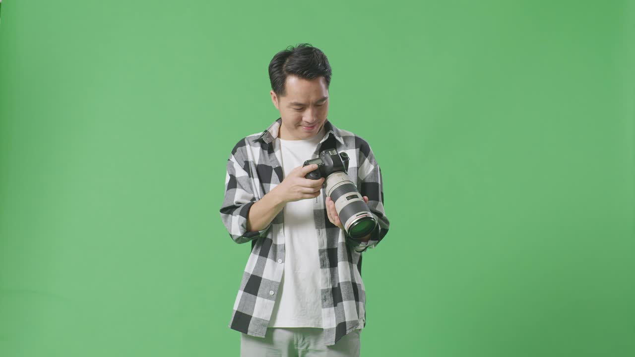 아시아 사진작가 는 스튜디오 의 녹색 스크린 배경 에 서 있는 동안 카메라 에 있는 사진 을 보고 미소 짓고 결과 에 만족 하고 있다