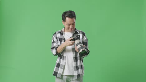 아시아 사진작가 는 스튜디오 의 녹색 스크린 배경 에 서 있는 동안 카메라 에 있는 사진 을 보고 미소 짓고 결과 에 만족 하고 있다