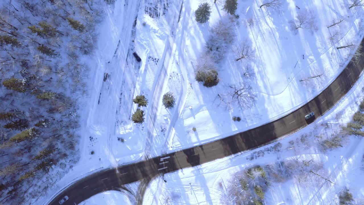 Aerial survey over snow covered curvy highway freeway inbetween a park with light traffic on beautiful winter day overlooking a frozen forest by the road 1-2
