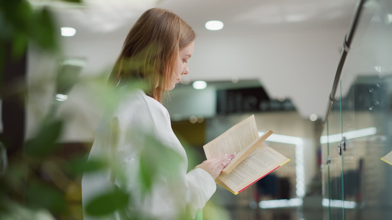 una mujer joven hojeando un libro mientras está sentada en un moderno centro comercial rodeada de luz suave, reflejos de vidrio y vegetación vibrante