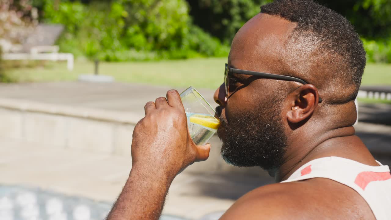 African American man smiling, sitting poolside with shades, holding glass, sipping lemonade