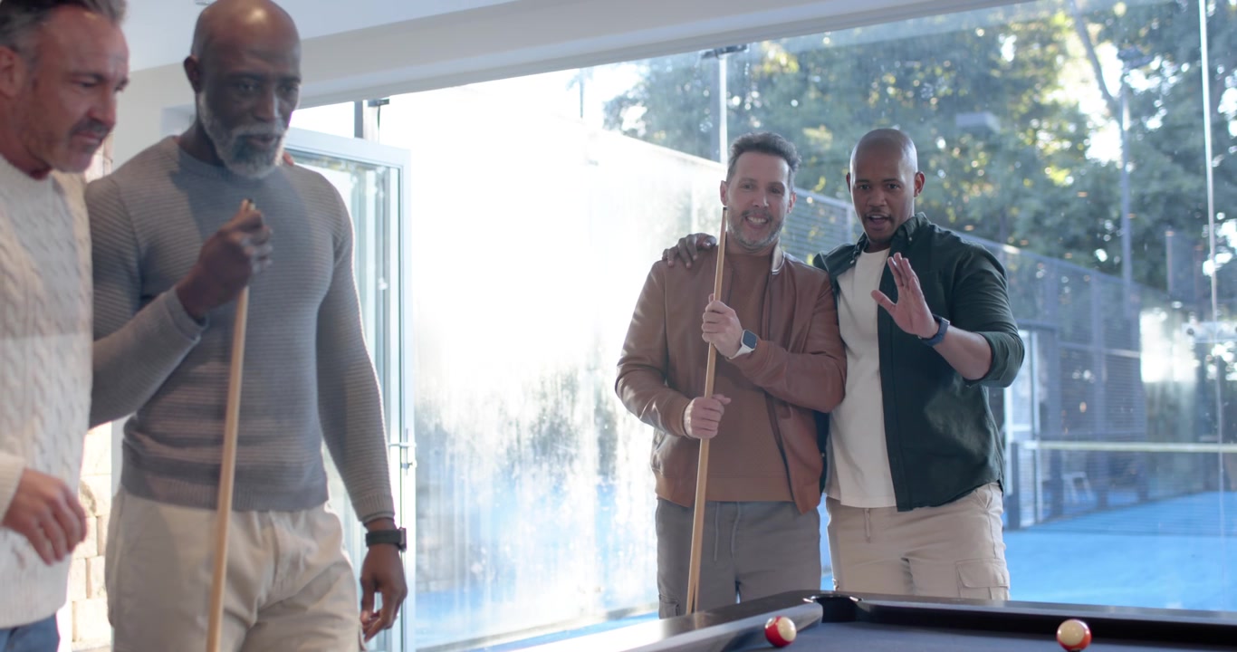 Diverse males with senior cheering after shot holding cues laughing watching balls blue pool table