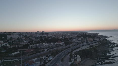 A-stunning-aerial-view-of-São-Pedro-do-Estoril-at-sunrise,-showcasing-the-moving-trains-and-the-beautiful-coastal-town-of-Cascais-in-Portugal