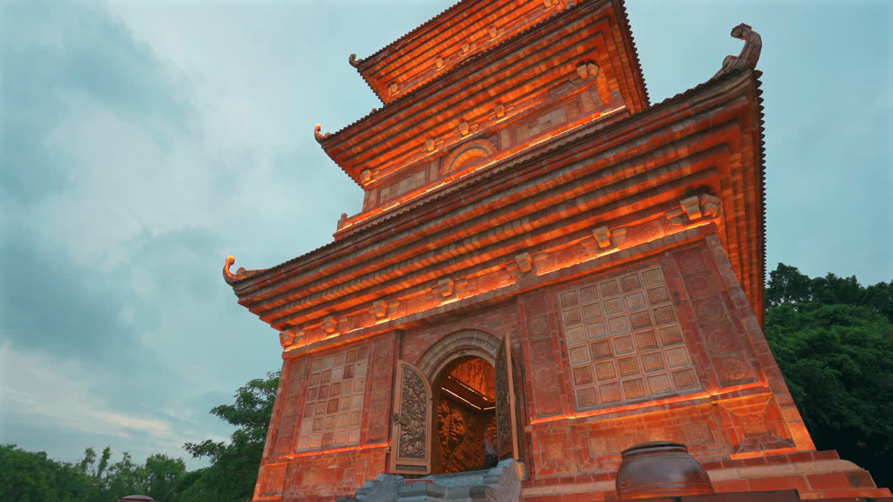 Toma en contrapicado de una majestuosa pagoda budista de varios niveles iluminada con cálidas luces naranjas contra un cielo crepuscular, con intrincadas tallas tradicionales y un visitante en la entrada en Ninh Binh