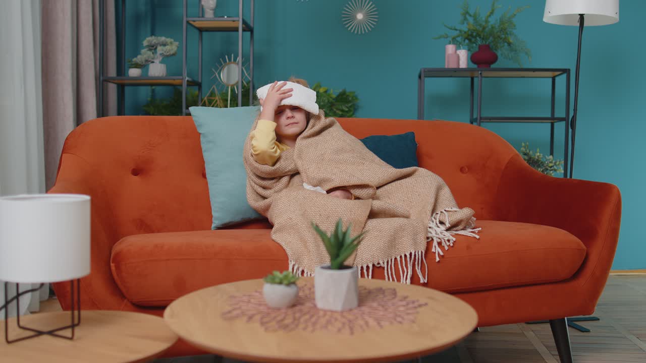 Child girl in plaid suffering from cold or allergy blows nose snot into napkin on sofa at home