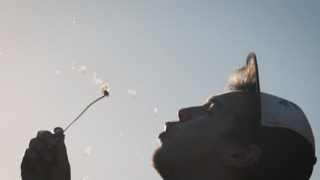 adult male wearing hat, blowing a dandelion at sunset in slow motion dreamy backlit close up golden hour