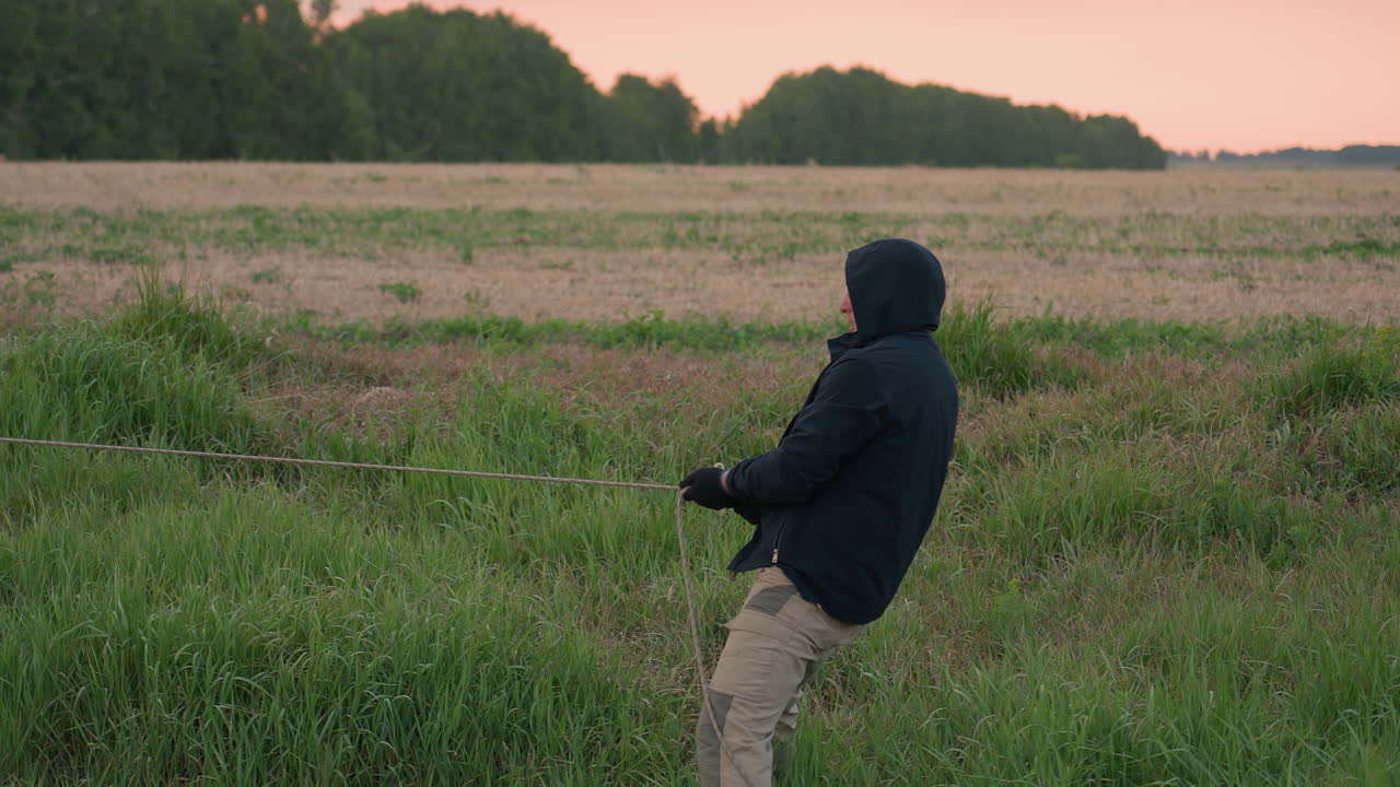 man in black hoodie dragging rope tied to deflated yellow balloon envelope through tall grass on rural farmland during serene sunset setup for hot air balloon pack up under pastel sky