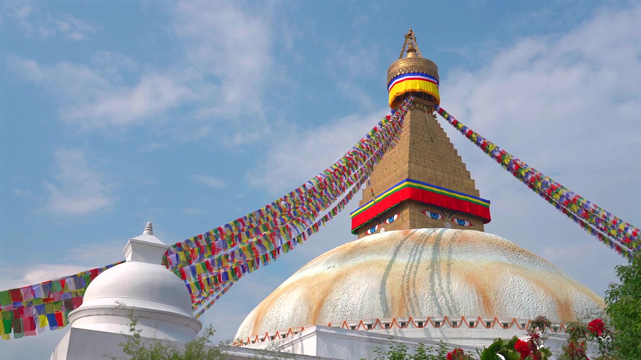 Un'ampia inquadratura statica che mostra il monumentale Stupa di Boudhanath accanto a un più piccolo santuario buddista bianco sotto un cielo azzurro brillante con nuvole leggere a Kathmandu, Nepal.