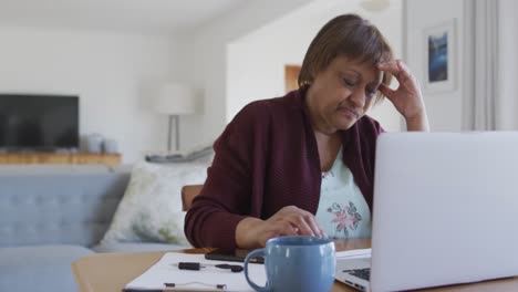 worried african american senior woman at dining table, using laptop and holding head