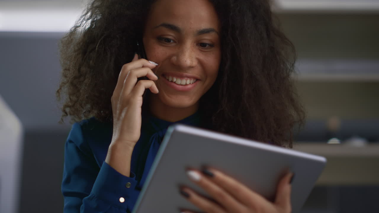 Smiling woman talking phone looking tablet pad in office. Business concept.