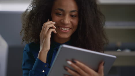 Smiling-woman-talking-phone-looking-tablet-pad-in-office.-Business-concept.